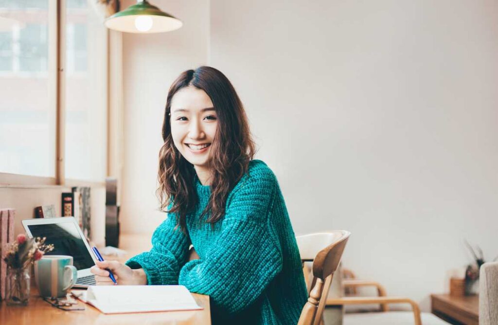 カフェで微笑みながら執筆する女性。AIを活用した創作活動が、より身近で楽しいものになる未来を象徴。